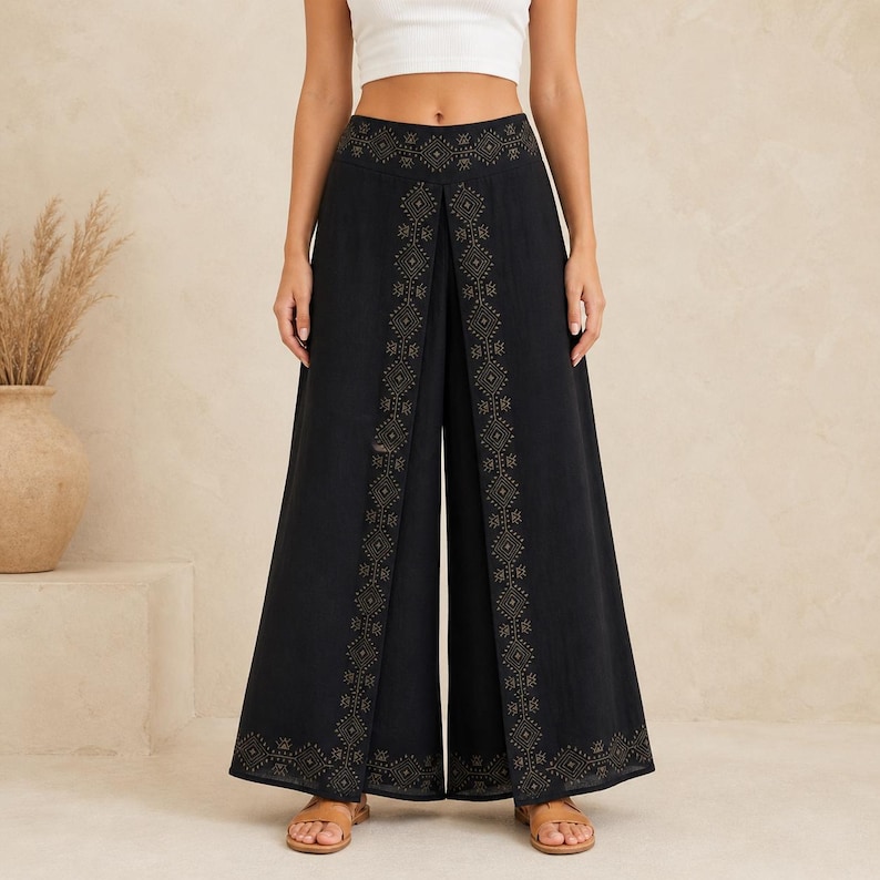 May include: Black palazzo pants with a split front and wide legs. The pants feature an intricate gold-colored geometric pattern along the split and waistband. The model is wearing a white crop top and sandals.