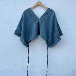 May include: A blue short-sleeved top featuring a deep V-neckline with black geometric embroidery. The garment has a loose, boxy fit with adjustable drawstring ties at the bottom hem on both sides, hanging from a wooden hanger against a white wall.