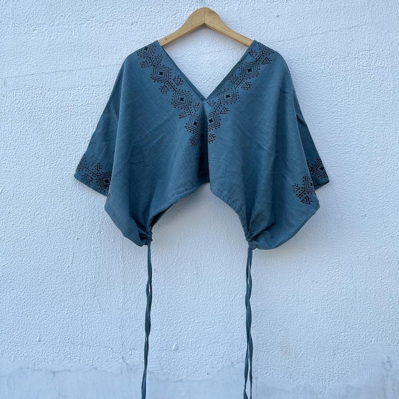 May include: A blue short-sleeved top featuring a deep V-neckline with black geometric embroidery. The garment has a loose, boxy fit with adjustable drawstring ties at the bottom hem on both sides, hanging from a wooden hanger against a white wall.