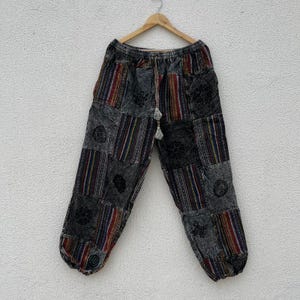 Puede incluir: Pantalones patchwork con una mezcla de rayas verticales grises, azules, rojas y amarillas y estampados florales. Los pantalones cuelgan de una percha de madera contra una pared blanca. Tienen cintura con cordón.