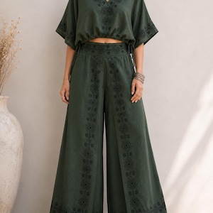 May include: A two-piece olive green outfit featuring a V-neck crop top with short sleeves and drawstring details, paired with wide-leg pants. Both pieces have black embroidered patterns. The outfit is made of a soft, flowing fabric.