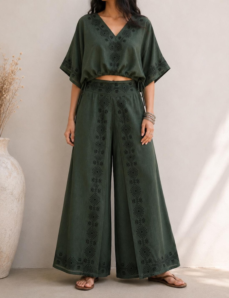 May include: A two-piece olive green outfit featuring a V-neck crop top with short sleeves and drawstring details, paired with wide-leg pants. Both pieces have black embroidered patterns. The outfit is made of a soft, flowing fabric.
