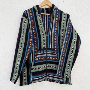 May include: A hooded jacket with a full zipper, featuring a striped pattern in black, blue, green, and red. The jacket has a front pocket and is hanging on a wooden hanger against a white background. The text "THE VIBES" is visible on the pocket.