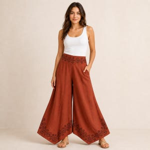 May include: A woman wearing a white tank top and rust-colored palazzo pants. The wide-leg pants feature a decorative black pattern along the waistband and hem. The pants have pockets.