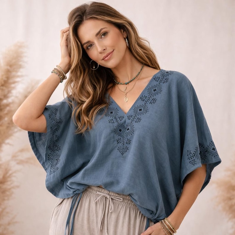 May include: A dusty blue V-neck top featuring short, wide sleeves and dark blue geometric embroidery along the neckline and sleeve edges. The garment has a relaxed, loose fit and is paired with light beige drawstring pants with a visible tie.