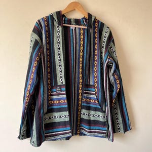May include: A hooded jacket with a colorful striped pattern in blue, green, red, and yellow. The jacket has a front zipper, two pockets, and a small patch with text. The jacket is hanging on a wooden hanger.