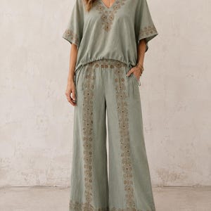 May include: Sage green two-piece outfit featuring a V-neck top and wide-leg pants. The top and pants have intricate embroidered detailing in a complementary color. The outfit is made of a lightweight fabric, perfect for warm weather.