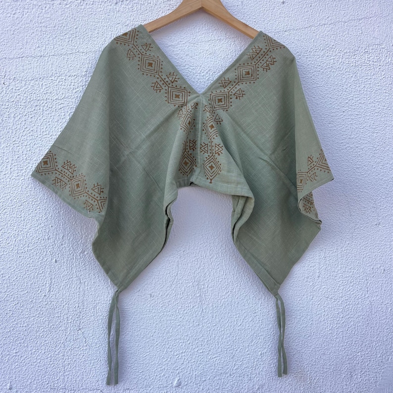 May include: A sage green poncho featuring a deep V-neckline and wide sleeves. The garment is decorated with brown geometric patterns along the neckline and sleeve edges, and includes two thin fabric ties hanging from the bottom corners.