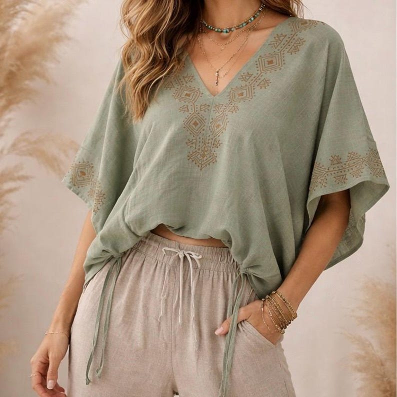May include: A sage green V-neck blouse featuring short, wide sleeves and a gold-toned geometric pattern around the neckline and sleeve edges. The blouse is paired with beige drawstring pants that have a visible tie at the center waist.