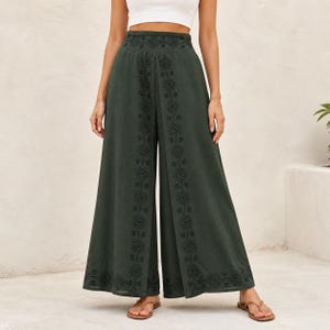 May include: Dark green palazzo pants with embroidered geometric patterns along the sides and waistband. The wide-leg pants are made of a flowing fabric and are worn with sandals. The pants have a high waist and a split front.