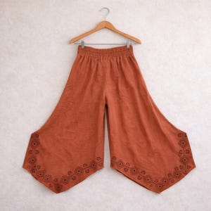 May include: Rust-colored palazzo pants with a wide leg design, hanging on a wooden hanger. The pants feature an elastic waistband and a decorative geometric pattern in a darker shade of brown along the bottom hem. The fabric appears to be lightweight.