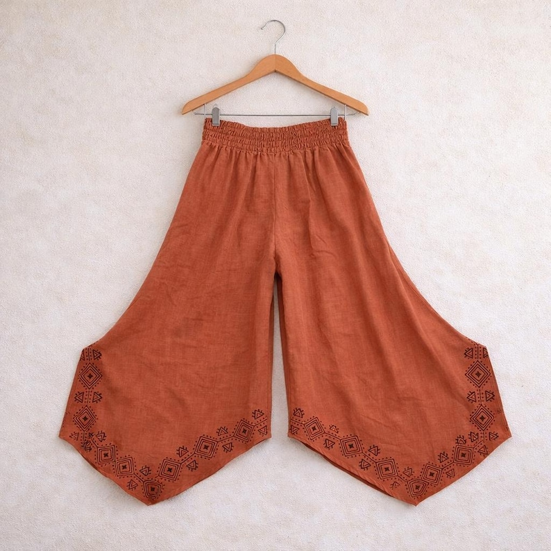 May include: Rust-colored palazzo pants with a wide leg design, hanging on a wooden hanger. The pants feature an elastic waistband and a decorative geometric pattern in a darker shade of brown along the bottom hem. The fabric appears to be lightweight.