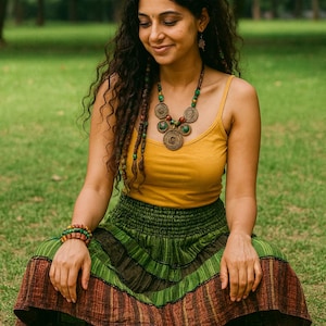 May include: A woman wearing a mustard yellow tank top, a green and brown striped skirt, and brown sandals sits on the grass. She is adorned with a beaded necklace and bracelet. The background is a blurred green park.
