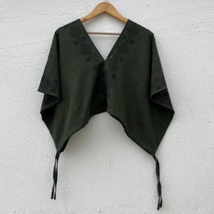 May include: A dark olive green poncho featuring a deep V-neckline and black geometric patterns printed along the collar and edges. The garment has a wide, draped silhouette with two long, thin fabric ties hanging from the bottom corners.