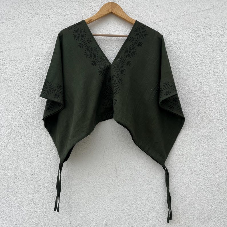 May include: A dark olive green poncho featuring a deep V-neckline and black geometric patterns printed along the collar and edges. The garment has a wide, draped silhouette with two long, thin fabric ties hanging from the bottom corners.