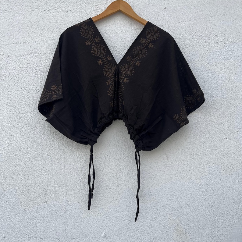 May include: A black short-sleeved top with a deep V-neckline and a drawstring hem featuring two hanging ties. The garment is decorated with a brown geometric pattern along the neckline and shoulders, creating a distinct contrast against the black fabric.