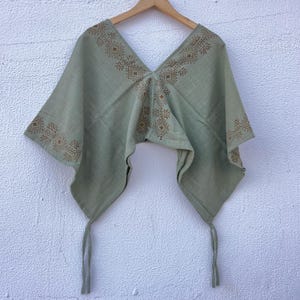 May include: A sage green poncho-style top with a V-neck and geometric brown embroidery. The lightweight fabric has a flowing design with tie closures at the bottom. The top is displayed on a wooden hanger against a white wall.