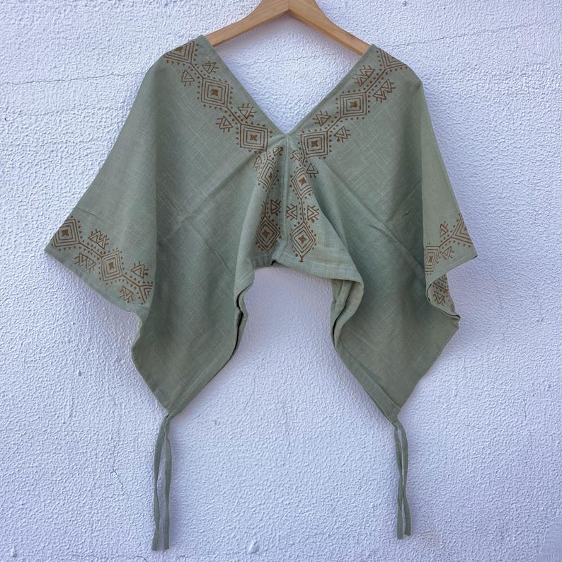 May include: A sage green poncho-style top with a V-neck and geometric brown embroidery. The lightweight fabric has a flowing design with tie closures at the bottom. The top is displayed on a wooden hanger against a white wall.