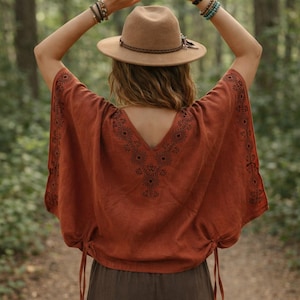 May include: A rust-colored short-sleeve top featuring a V-neckline and black geometric embroidery along the neckline and sleeve edges. The garment includes side drawstrings at the hem and is paired with a tan fedora hat with a braided band.