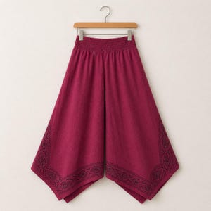 May include: A pair of burgundy palazzo pants hanging on a wooden hanger. The pants have a wide leg design with an elastic waistband. The bottom edges feature a black geometric pattern.