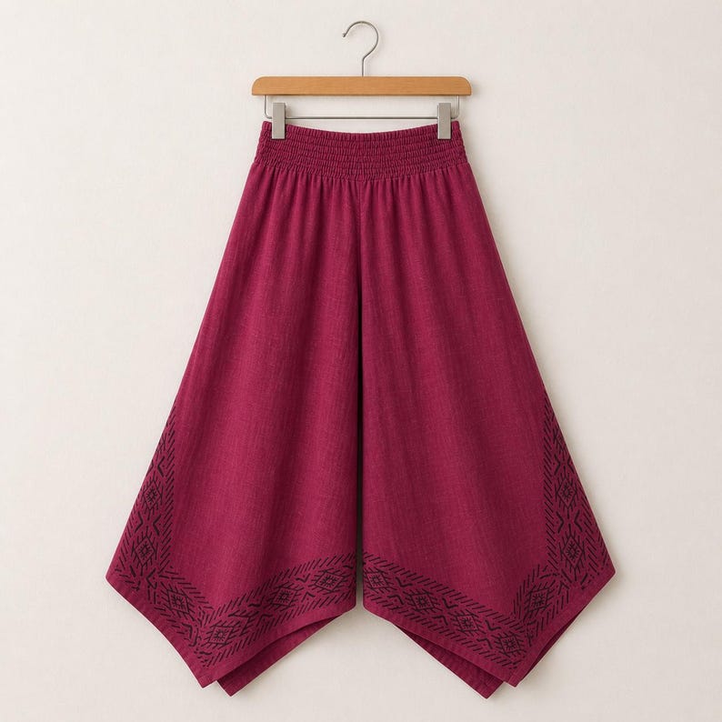 May include: A pair of burgundy palazzo pants hanging on a wooden hanger. The pants have a wide leg design with an elastic waistband. The bottom edges feature a black geometric pattern.