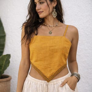Pode incluir: Um top curto amarelo-mostarda estilo lenço com alças finas e um padrão de chevron castanho escuro ao longo da bainha inferior. A peça tem uma bainha frontal pontiaguda e é combinada com um colar de contas em camadas e brincos.