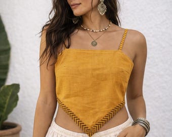 May include: A mustard yellow handkerchief-style crop top featuring thin shoulder straps and a dark brown chevron pattern along the bottom hem. The garment has a pointed front hemline and is paired with a layered beaded necklace and earrings.