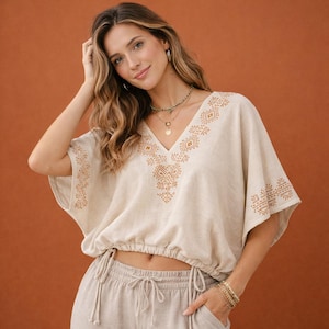 May include: A beige cropped top with short, wide sleeves and a V-neckline. The garment features brown geometric embroidery around the neckline and sleeve edges. It has an elasticized hem with adjustable drawstring ties at the sides.