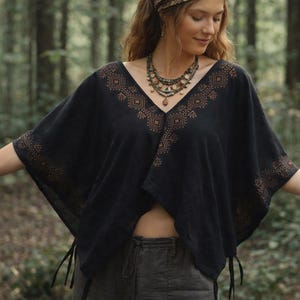 May include: A black V-neck poncho featuring a copper-colored geometric pattern along the neckline and sleeve edges. The garment has a loose, draped fit with side ties at the hem, paired with a multi-layered beaded necklace with earth-toned stones.