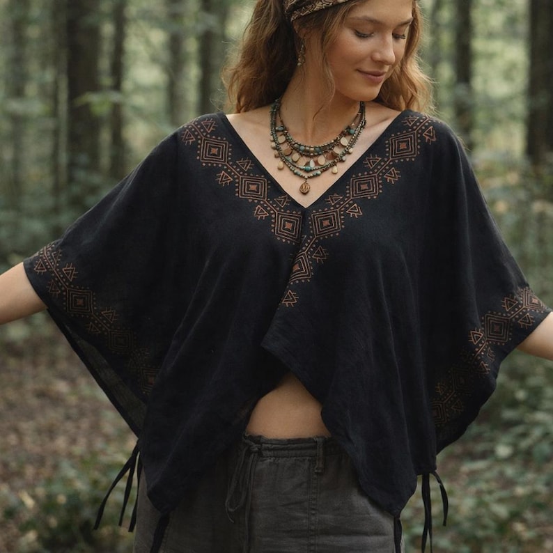 May include: A black V-neck poncho featuring a copper-colored geometric pattern along the neckline and sleeve edges. The garment has a loose, draped fit with side ties at the hem, paired with a multi-layered beaded necklace with earth-toned stones.