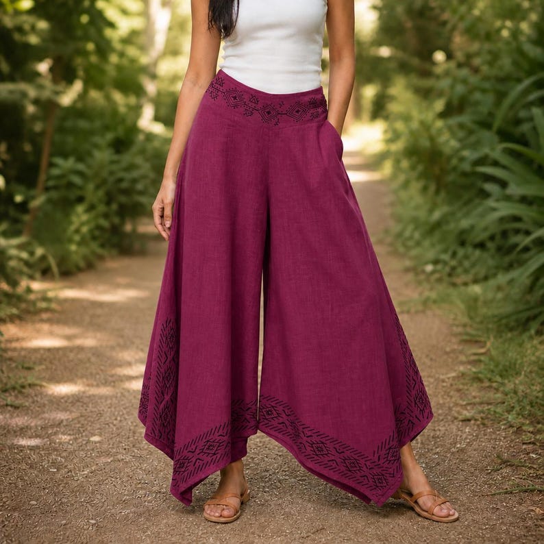 May include: Wide-leg, burgundy pants with a decorative embroidered waistband and hem. The pants have pockets and a flowing design. The model is wearing a white top and brown sandals. The pants are made of a lightweight fabric.