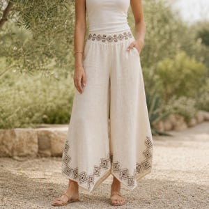 May include: Cream-colored wide-leg pants with a geometric embroidered design at the waist and hem. The pants are made of a lightweight fabric and feature pockets. The model is wearing a white tank top and sandals.