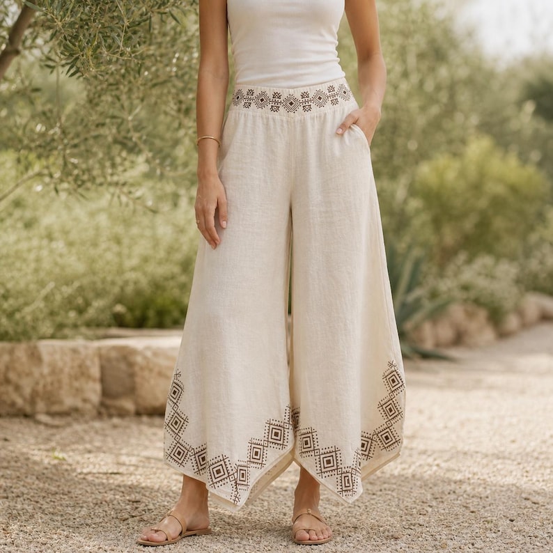 May include: Cream-colored wide-leg pants with a geometric embroidered design at the waist and hem. The pants are made of a lightweight fabric and feature pockets. The model is wearing a white tank top and sandals.