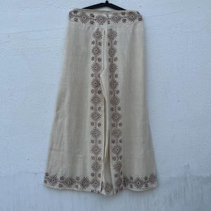 May include: Cream-colored wide-leg pants with a geometric pattern in brown. The pants are hanging on a black hanger against a neutral background. The decorative pattern runs along the sides and the bottom of the pants.