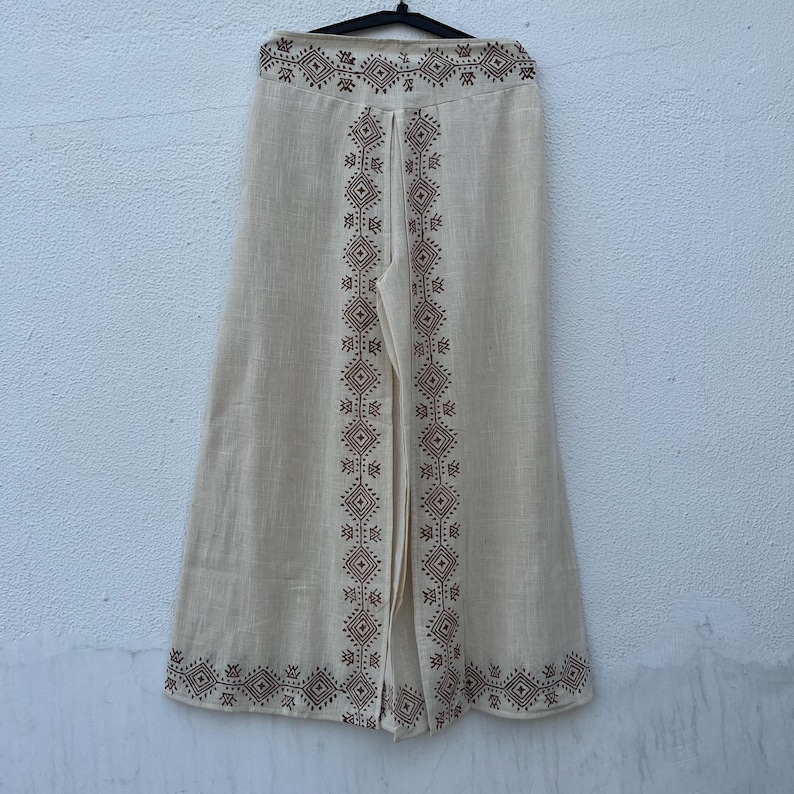 May include: Cream-colored wide-leg pants with a geometric pattern in brown. The pants are hanging on a black hanger against a neutral background. The decorative pattern runs along the sides and the bottom of the pants.