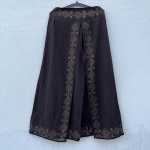May include: Dark brown wide-leg pants with a decorative geometric pattern in a lighter brown color. The pants are hanging on a black hanger against a white wall. The decorative pattern is along the sides and the top of the pants.