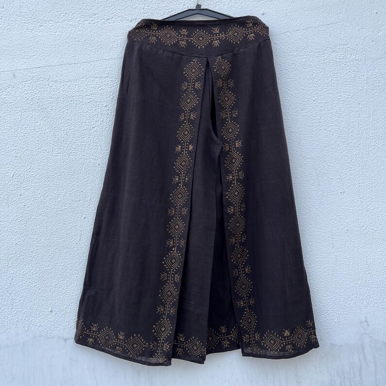 May include: Dark brown wide-leg pants with a decorative geometric pattern in a lighter brown color. The pants are hanging on a black hanger against a white wall. The decorative pattern is along the sides and the top of the pants.