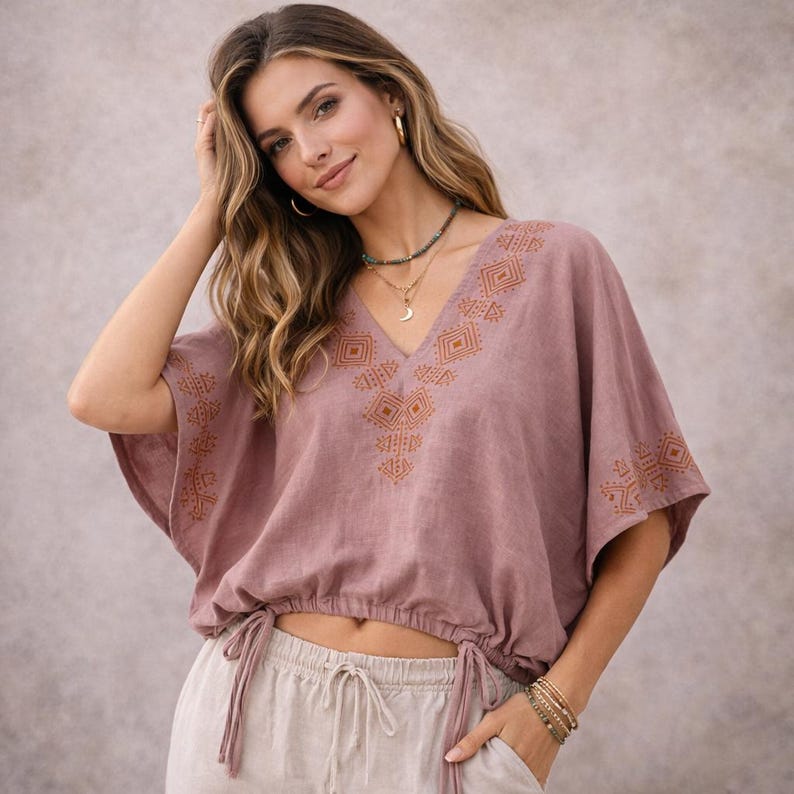 May include: A dusty rose-colored short-sleeve blouse featuring a V-neckline with intricate brown geometric embroidery. The garment has a cinched hemline with adjustable drawstring ties at the sides and loose, wide sleeves, crafted from a textured fabric.