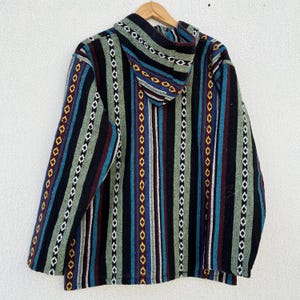 May include: A hooded jacket with a striped pattern in black, green, blue, and maroon. The jacket is hanging on a wooden hanger against a white wall. The stripes are vertical and feature geometric shapes.