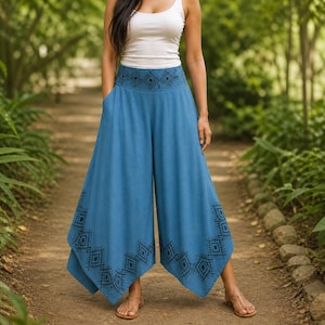 May include: A woman wearing a white tank top and blue palazzo pants. The pants have a wide leg design with black geometric patterns along the waistband and hem. The pants have pockets and are made of a lightweight fabric.