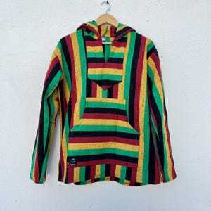 Può includere: Una felpa con cappuccio a righe rosse, gialle, verdi e nere. La felpa con cappuccio ha una tasca a marsupio e un cappuccio. Il testo "THE GOOD VIBES" è visibile in basso a sinistra della felpa con cappuccio.