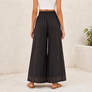 May include: Black wide-leg pants with an elastic waistband and a decorative pattern near the hem. The pants are made of a dark fabric and are paired with sandals. The pants are a casual style.