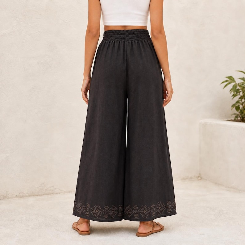 May include: Black wide-leg pants with an elastic waistband and a decorative pattern near the hem. The pants are made of a dark fabric and are paired with sandals. The pants are a casual style.
