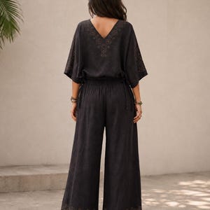 May include: A black two-piece outfit featuring a V-neck top with embroidered details and wide-leg pants. The top has short sleeves and a drawstring waist. The pants have an embroidered design at the hem. The outfit is made of a soft, flowing fabric.