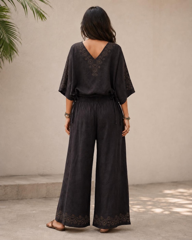 May include: A black two-piece outfit featuring a V-neck top with embroidered details and wide-leg pants. The top has short sleeves and a drawstring waist. The pants have an embroidered design at the hem. The outfit is made of a soft, flowing fabric.