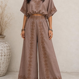 May include: A two-piece taupe outfit featuring a V-neck top and wide-leg pants. The top has short sleeves and a drawstring waist. Both pieces are adorned with intricate brown embroidered geometric patterns. The outfit is made of a soft, flowing fabric.