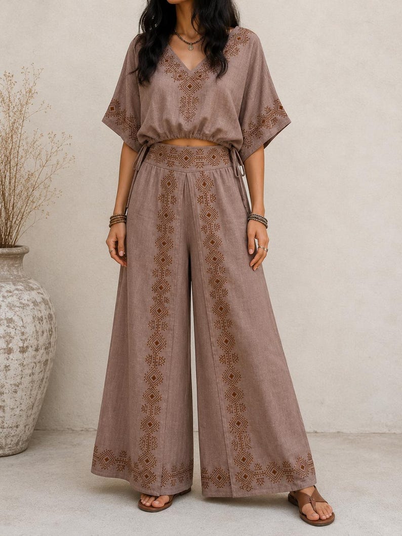 May include: A two-piece taupe outfit featuring a V-neck top and wide-leg pants. The top has short sleeves and a drawstring waist. Both pieces are adorned with intricate brown embroidered geometric patterns. The outfit is made of a soft, flowing fabric.