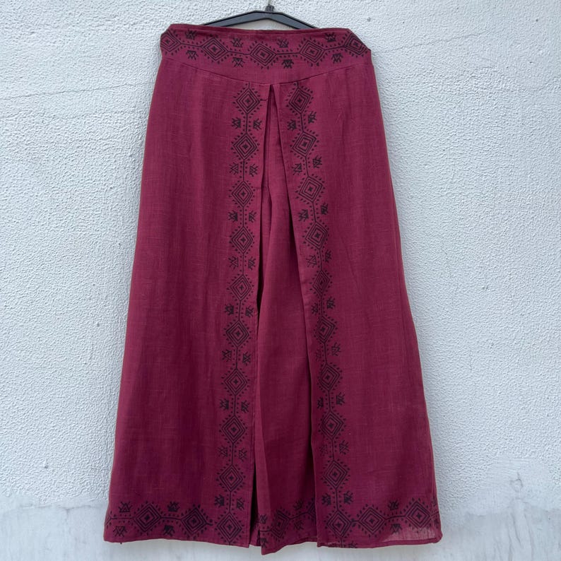 May include: Burgundy-colored wide-leg pants with a decorative geometric pattern in black. The pants are hanging on a black hanger. The fabric appears to be linen or a similar lightweight material.