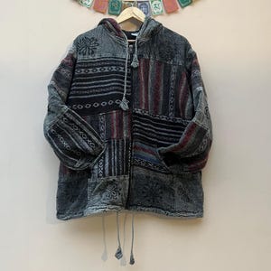 Puede incluir: Chaqueta con capucha de patchwork en tonos grises, negros y burdeos. La chaqueta presenta cierre de cremallera, bolsillos delanteros y capucha con cordón. El diseño incorpora patrones florales y geométricos, creando una apariencia única y texturizada. La chaqueta está colgada en una percha de madera.