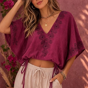 May include: A deep magenta cropped blouse featuring a V-neckline and wide short sleeves, both adorned with black geometric embroidery. The hem includes an adjustable drawstring with long ties on each side, paired with light beige drawstring pants.